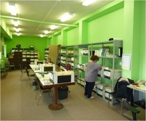 Une employée cherche des documents à ScanWerks pour les entrer dans la base de données centrale. Dans la pièce, il y a des étagères avec des livres et des papiers. La pièce est spacieuse et claire, les murs sont vert lime.