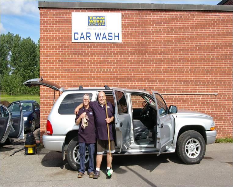 Deux membres du personnel de Waterwerks, tenant un chiffon et une vadrouille, devant une voiture fraîchement lavée aux portes ouvertes. Le lave-auto WaterWerks